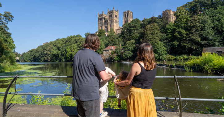 family with back to camera stand and admire the view of Durham Cathedral from the banks of the River Wear on a bright sunny day